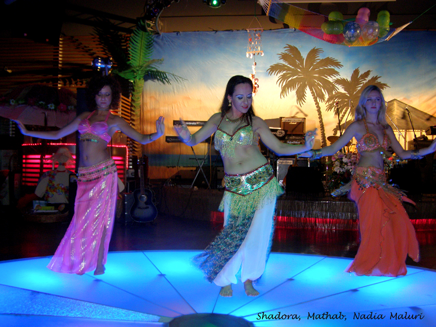 Trzy tancerki brzucha w kostiumach scenicznych prezentują układ taneczny na podświetlanej scenie w Łanach, z dekoracjami tropikalnymi w tle.